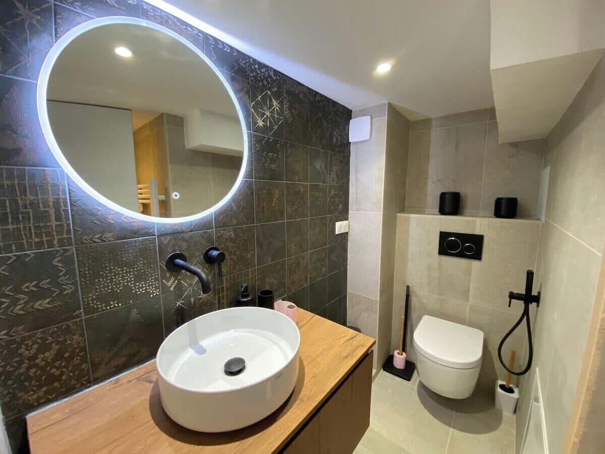 A modern bathroom with a round mirror, a white sink, a toilet, and a shower area.