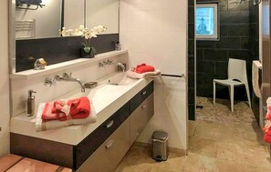 Une salle de bain moderne avec un grand miroir, un lavabo blanc et une serviette rouge. On peut voir une poubelle et une chaise blanche.