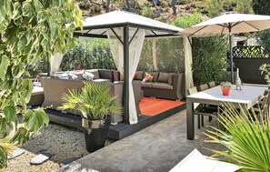 Un patio couvert avec un espace salon extérieur comprenant un canapé et des fauteuils, entouré de plantes en pot et agrémenté d’une table avec un parasol.