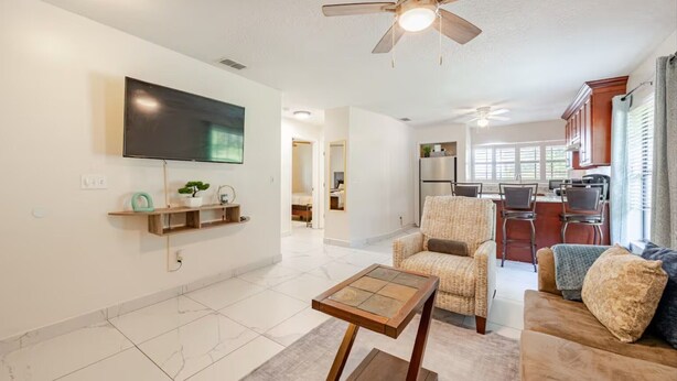 A modern living room with a flat-screen TV, a ceiling fan, a dining area with a kitchen in the background, and a hallway leading to another room.