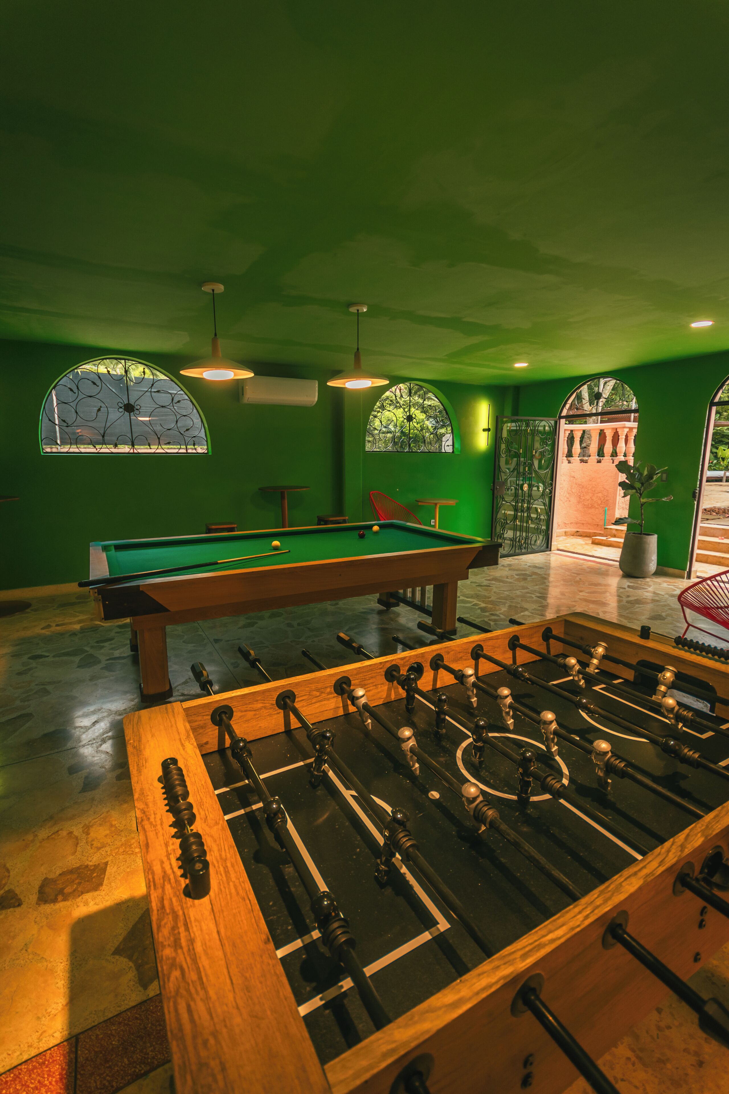 A pool table, foosball table, and a billiards table in a room with green walls and large windows.