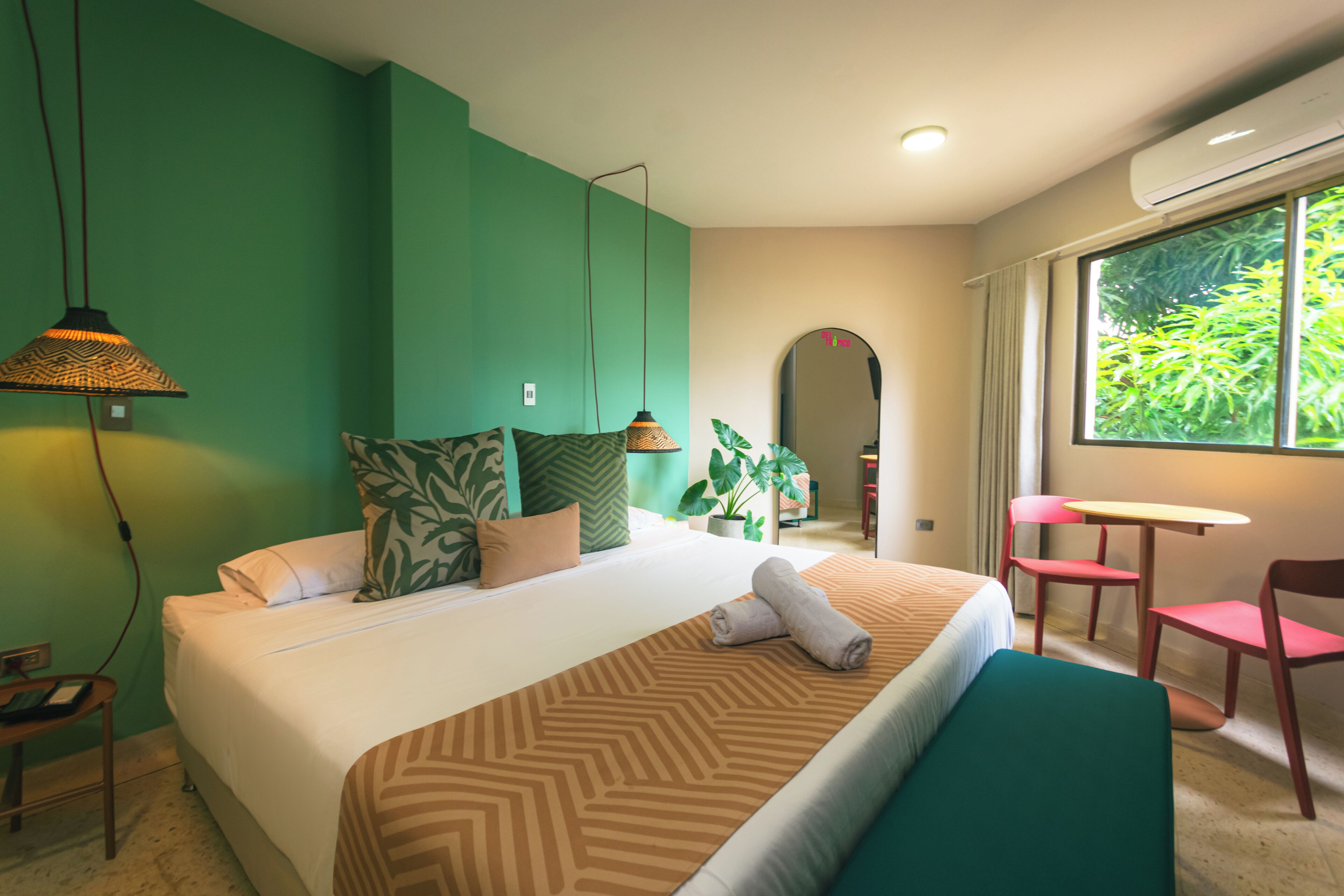 A hotel room with a bed, a table, chairs, a potted plant, and a window with a view.