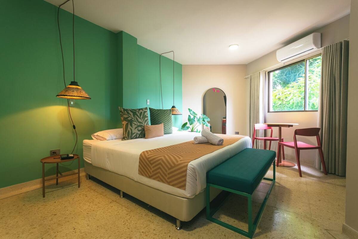 A modern hotel room with a bed, bedside table, lamp, bench, and a view of greenery through the window.
