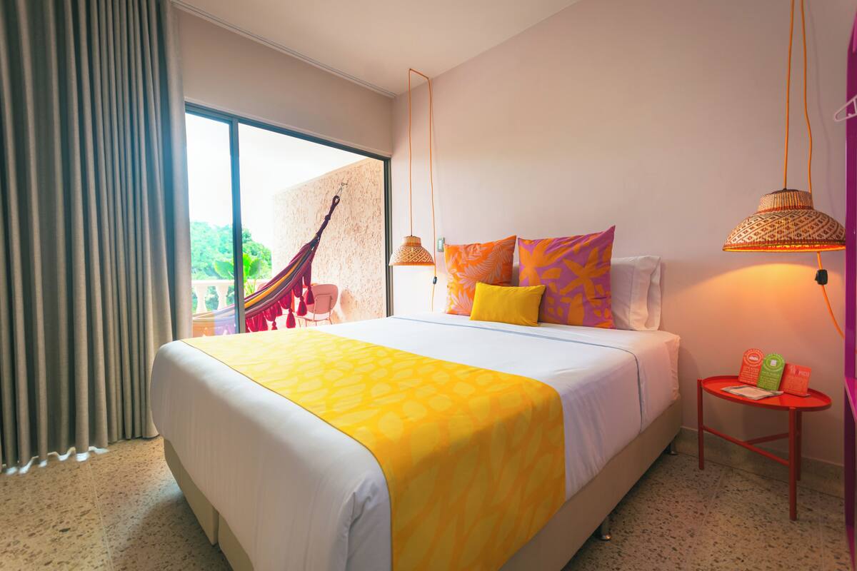 A hotel room with a large bed, a hammock, a small table with books, and a view of greenery outside.