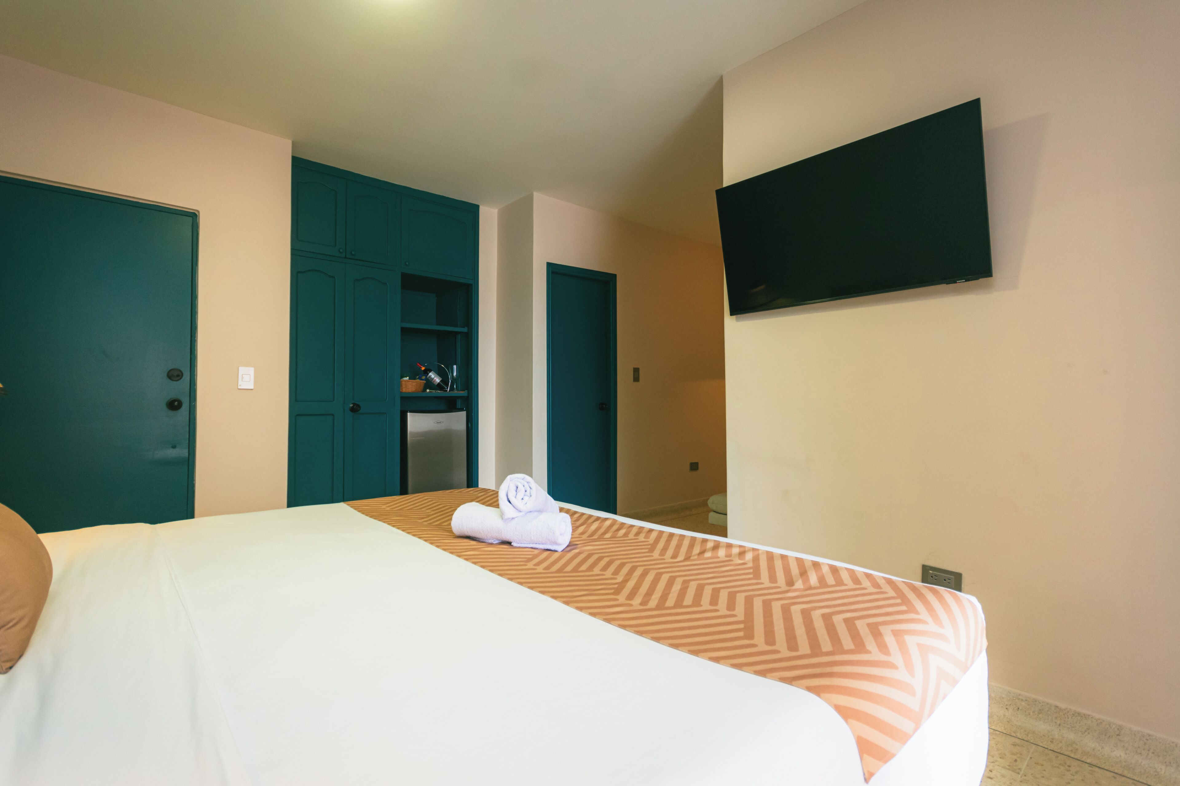 A hotel room with a bed, a TV mounted on the wall, and teal-colored doors.