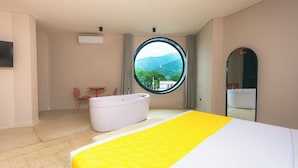 A hotel room with a bed, a round window with a mountain view, a white bathtub, and a small table with a chair. - Del Tropico Hotel (Sopetrán)