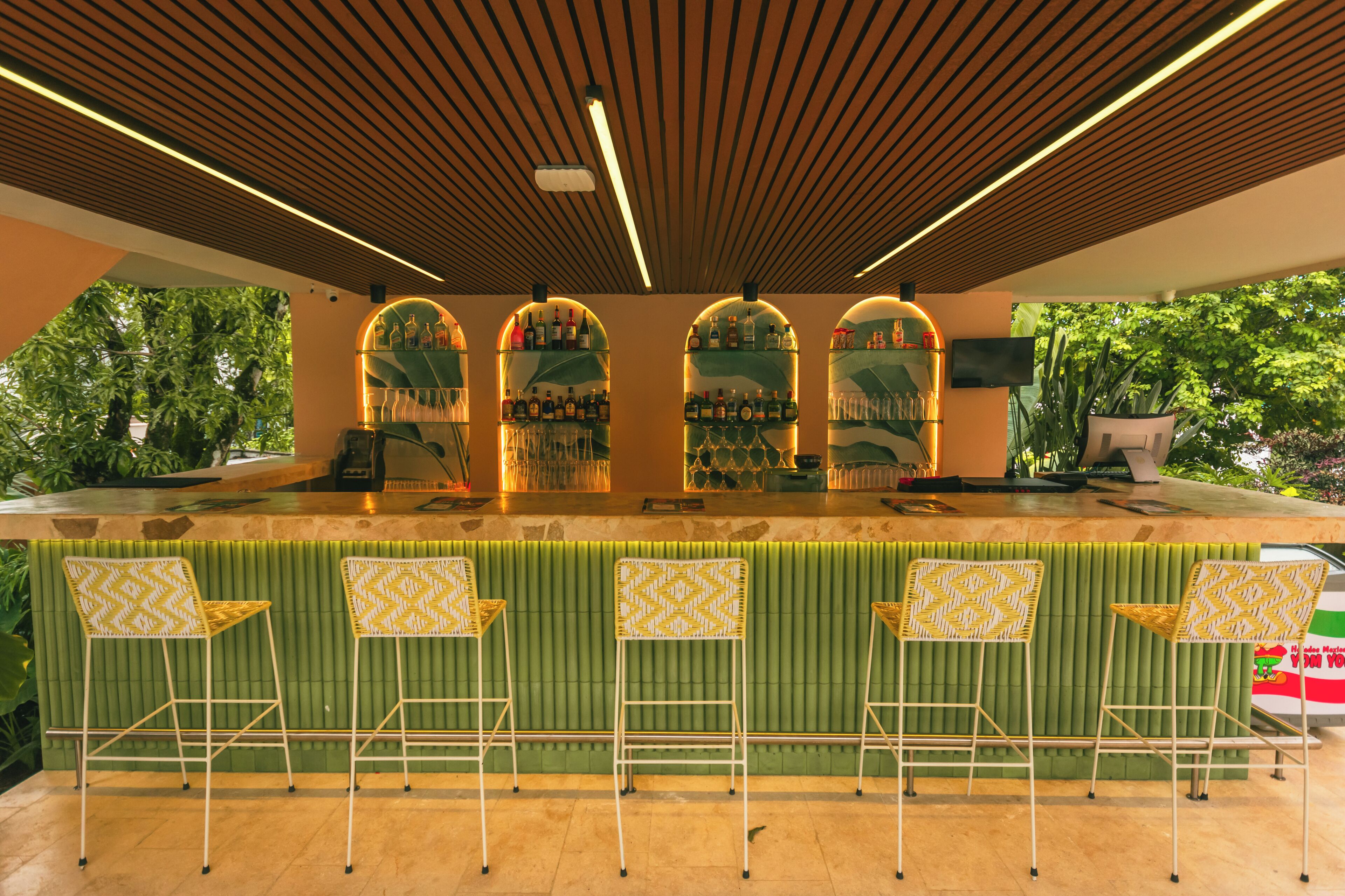 Un espace bar doté d’un banc rembourré de couleur verte, d’un plafond en bois avec des luminaires linéaires et d’une vue sur la verdure à l’extérieur.