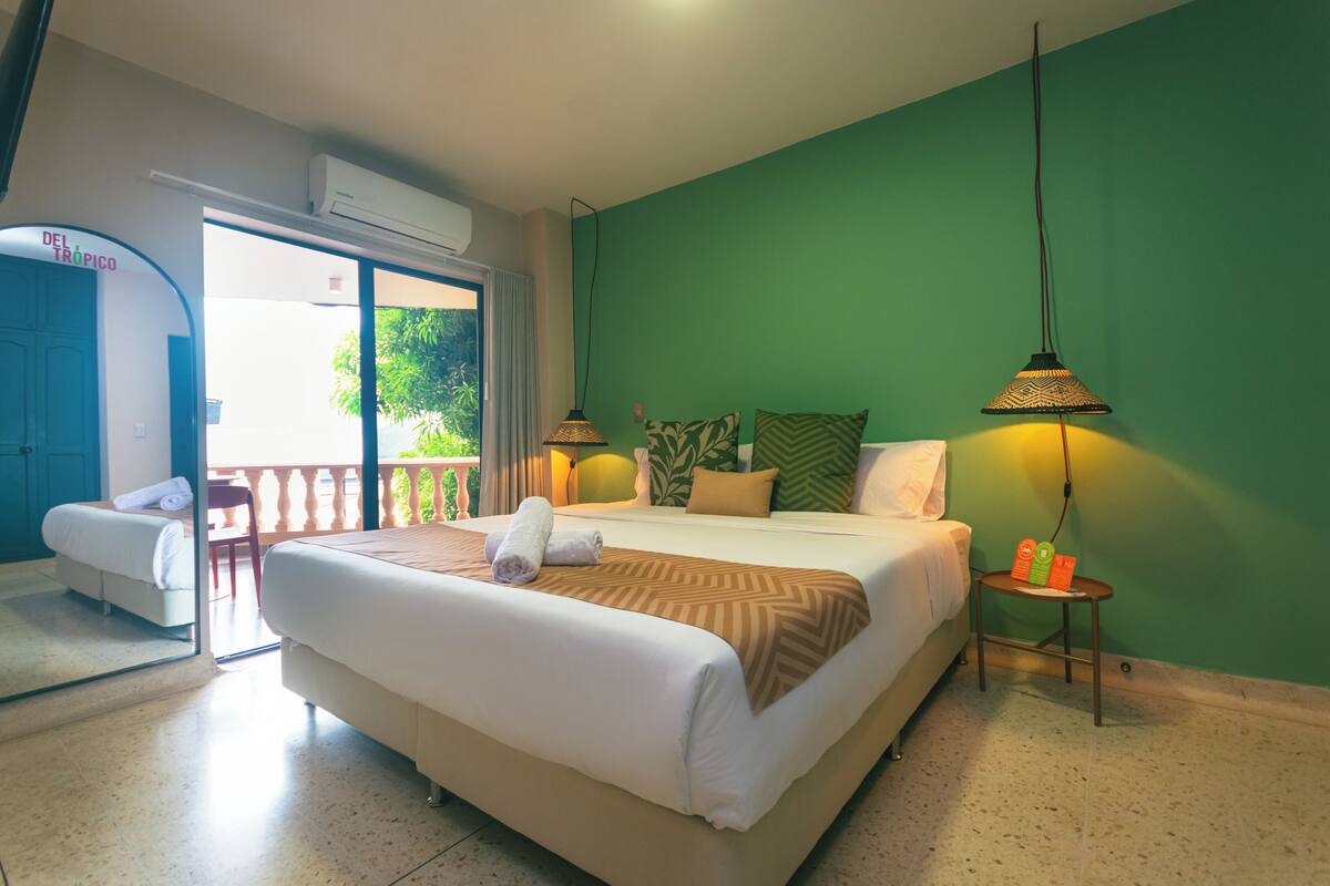 A hotel room with a bed, two bedside lamps, a small table with toiletries, a chair, and a balcony with a view.