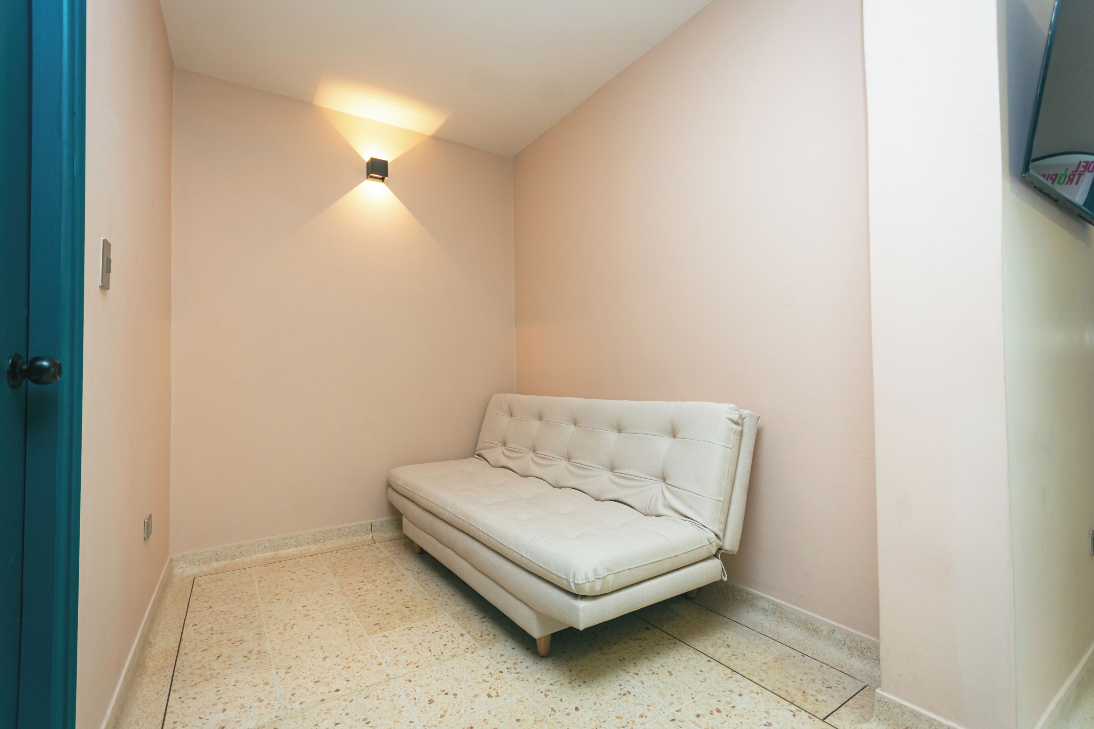 A small, narrow room with a single white sofa, a wall-mounted light, and a door.