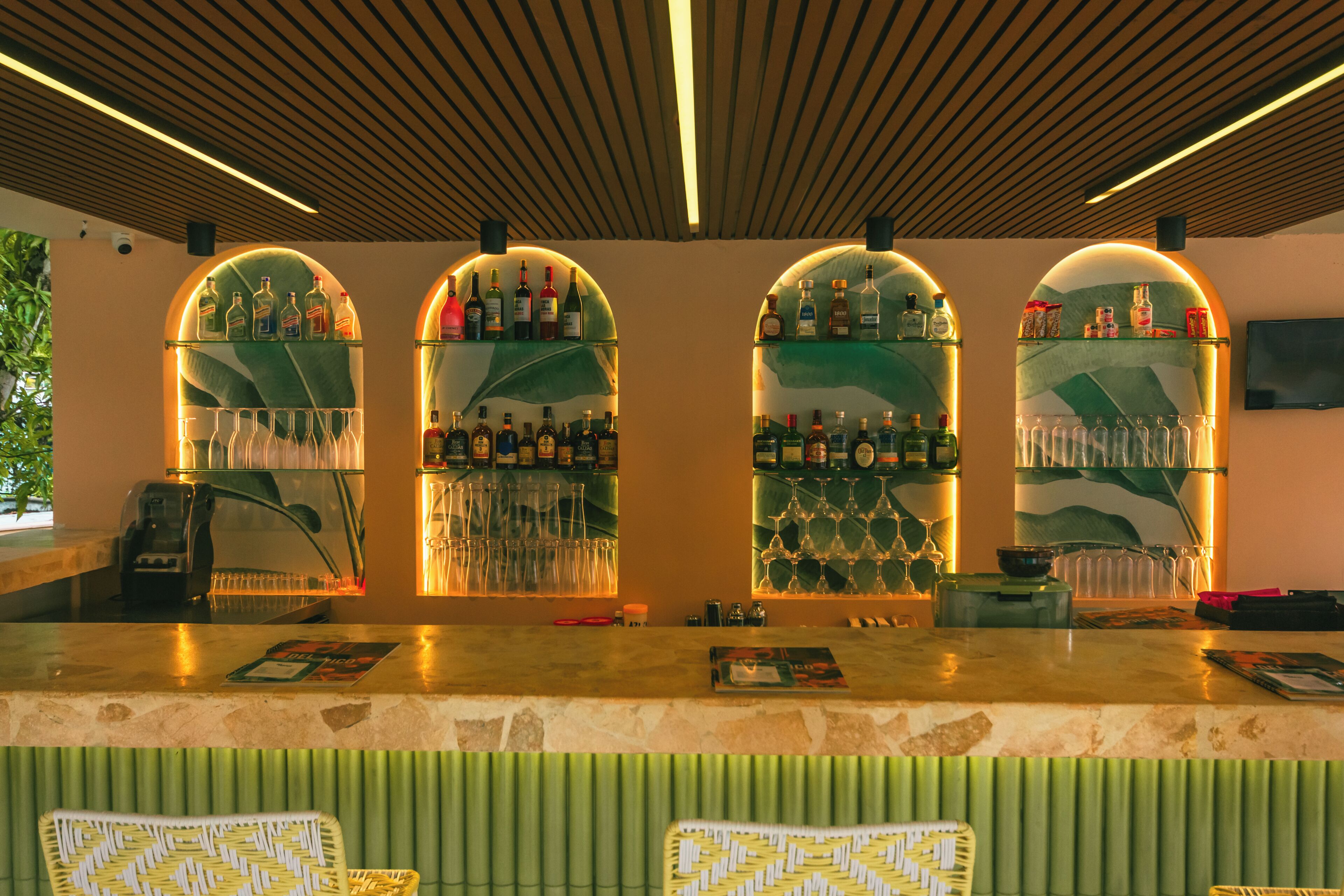 A bar area with arched shelves displaying bottles and glasses, a countertop with a menu, and a green upholstered chair.