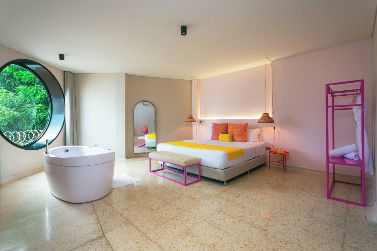 A modern bedroom with a round window, a large bed with a yellow and orange bedspread, a white bedside table, a pink shelf, and a round bathtub.
