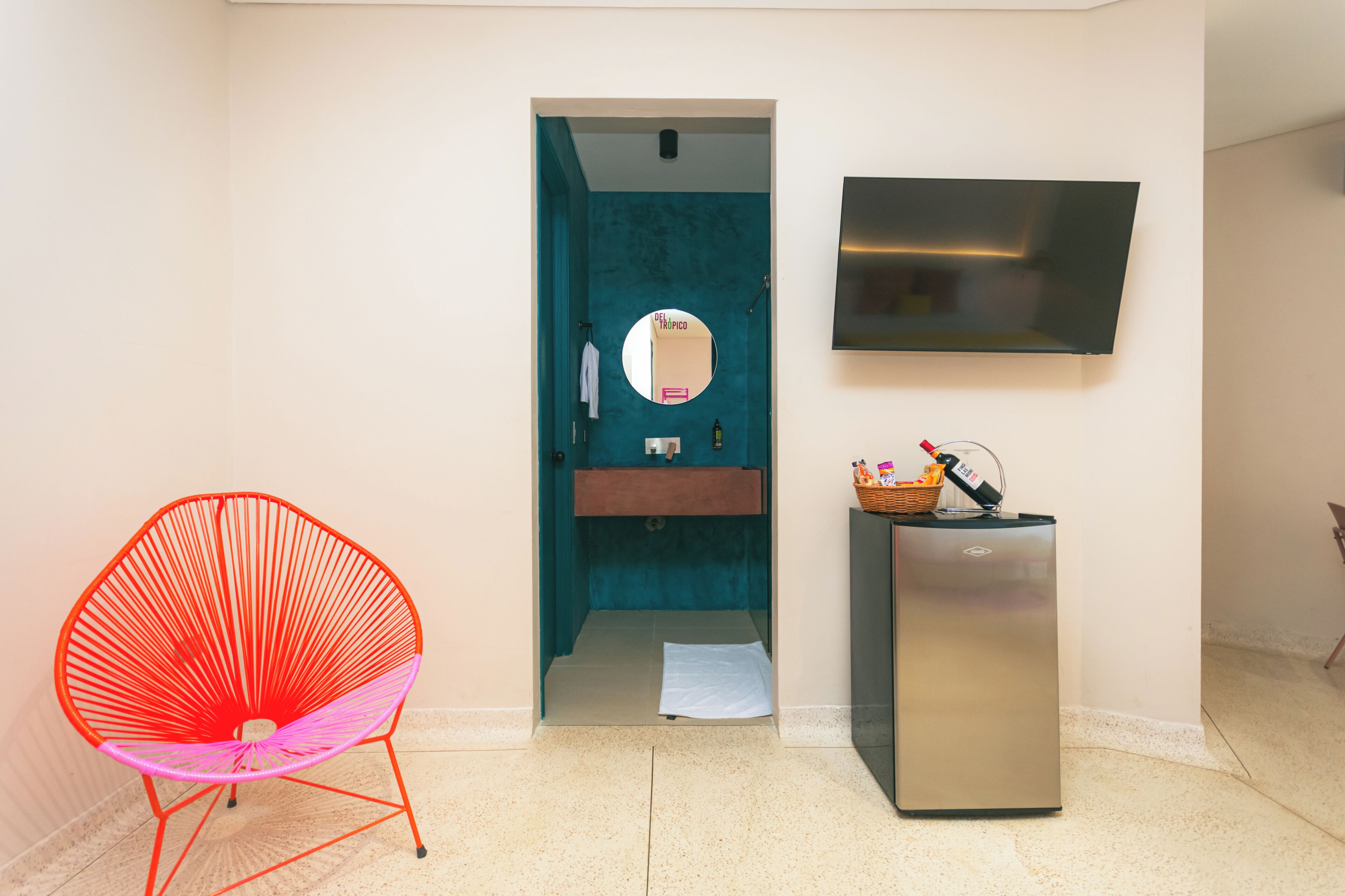 A room with a teal-colored wall, a flat-screen TV mounted above a sink, a red chair, and a refrigerator.