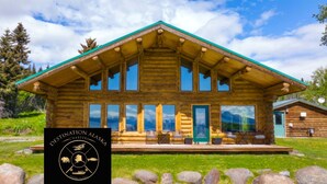 A log cabin with a green roof and large windows, surrounded by a grassy area and rocks. - Destination Alaska Charters - Cabin 1 (Homer)