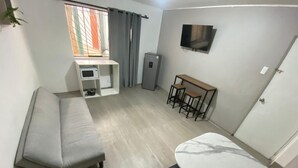 Quarto compacto com sofá, uma pequena mesa com cadeiras, televisão na parede e um refrigerador.