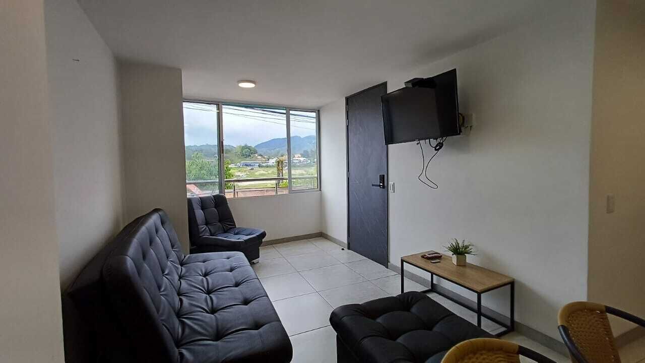 A compact living space with a sofa, armchair, and a small table. A TV is mounted on the wall, and there's a window offering a view of greenery and hills.