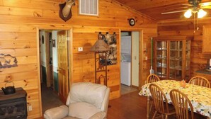 L’intérieur d’un chalet en bois, avec une tête de cerf accrochée au mur, une table à manger entourée de chaises, une cheminée et un ventilateur de plafond.