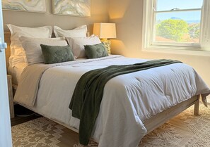 A neatly made bed with a patterned blanket and pillows, a bedside lamp, and a view of a tree through a window.
