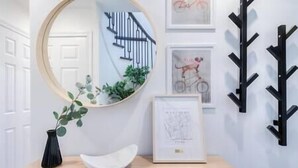 Une pièce avec un miroir rond, un vase blanc contenant des feuilles vertes, un bol blanc et des œuvres d’art encadrées accrochées au mur.