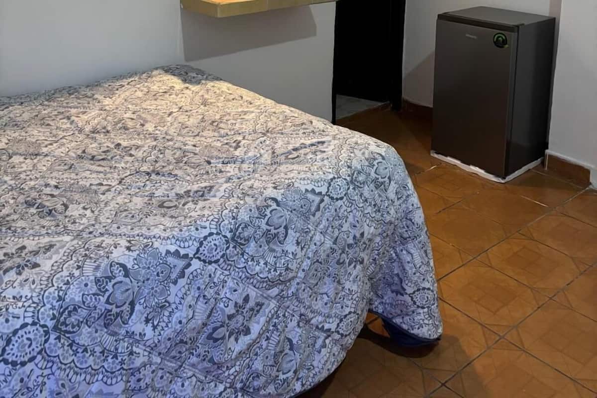 A bedroom with a patterned bedspread, a bedside table with a mirror and lights, a small refrigerator, and a door.