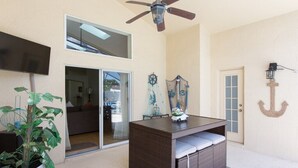 Una habitación con ventilador de techo, televisor montado en la pared, una planta grande en maceta, una mesita de mimbre con cojines y una puerta con panel de vidrio.