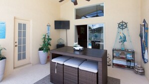 Un patio techado con un ventilador de techo, una mesita con un objeto decorativo, un banco con cojines, una planta en maceta y un letrero de bienvenida.