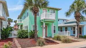 A two-story house with a balcony, surrounded by palm trees and a white picket fence. - Seashell Escape--3 BR House in Village Walk, Sleeps 10, Near Beach, Dogs OK! (Port Aransas)