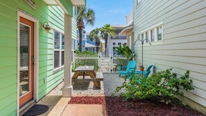 A narrow, green-sided building with a white door, a small patio with a table and chairs, and a white picket fence. - Seashell Escape--3 BR House in Village Walk, Sleeps 10, Near Beach, Dogs OK! (Port Aransas)