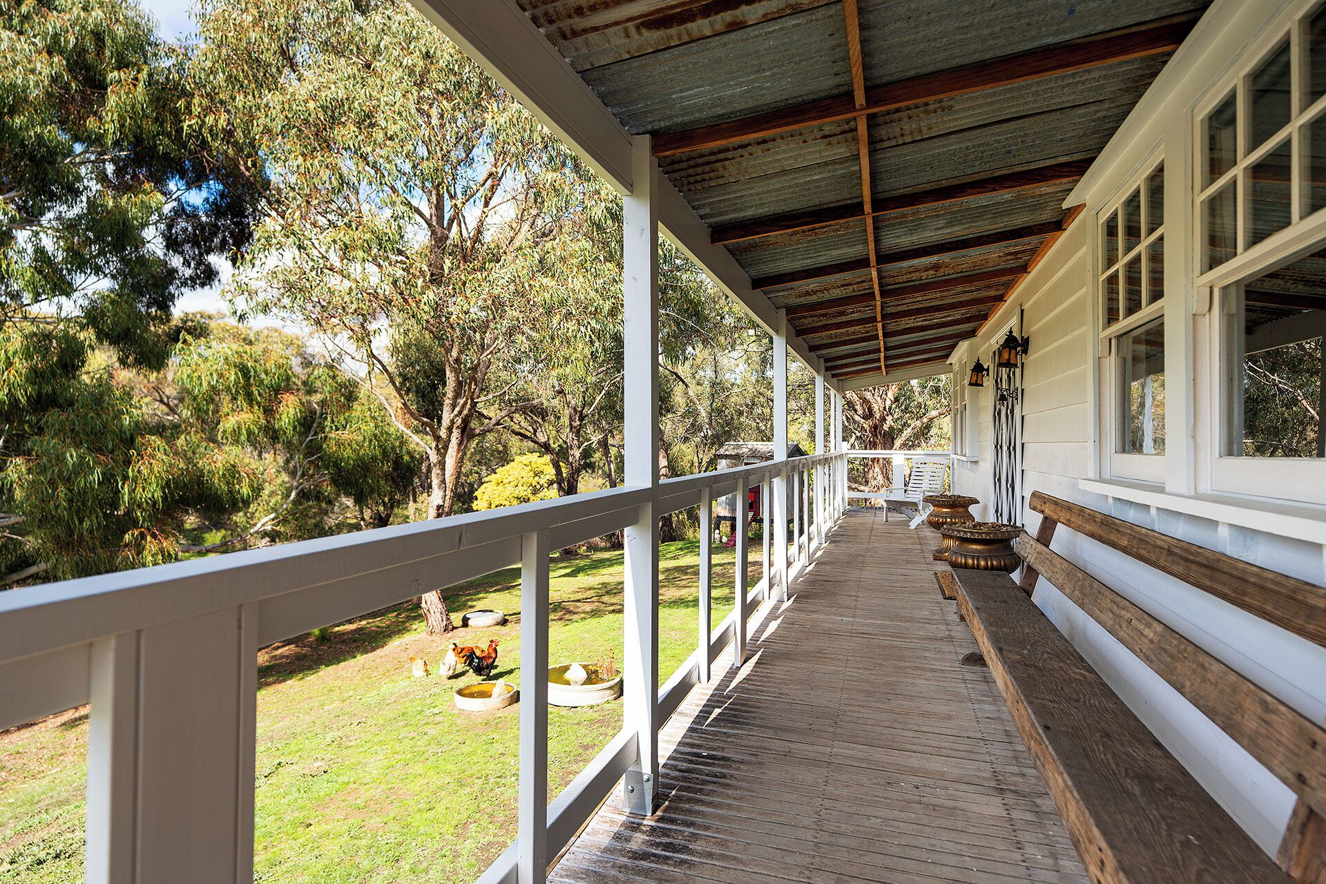 Une véranda couverte avec un plancher en bois, des balustrades blanches et une vue sur un jardin où l’on peut voir des poulets et des arbres.