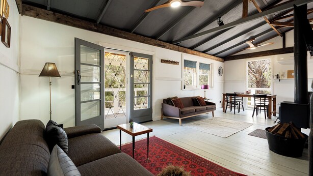 Un vaste espace de vie comprenant un canapé, une petite table et un tapis rouge. Le plafond est orné de poutres en bois et une grande fenêtre offre une vue sur les arbres.