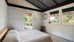 Une chambre avec un grand lit, deux fenêtres avec volets, un plafond en bois et une vue sur l’extérieur.