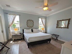 A bedroom with a bed, a ceiling fan, two bedside lamps, a window with curtains, a wall mirror, and a small dresser. - Hibiscus Hideaway: Pool, Spa & Steps to the beach (Cocoa Beach)