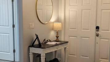 A hallway with a white door, a mirror, a table with a lamp, and a bench.