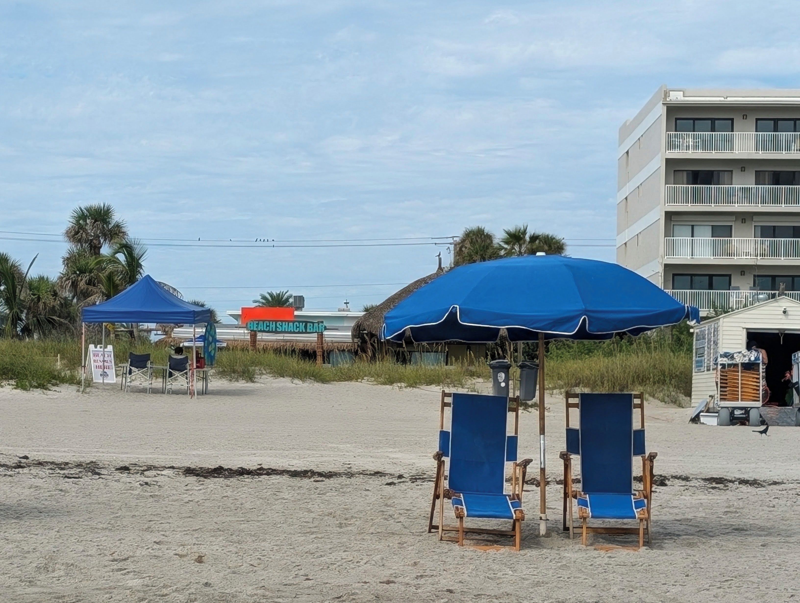 Una playa con dos sillas de playa azules bajo una gran sombrilla azul, un bar tipo choza en la playa y un edificio de varios pisos al fondo.