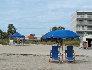 Una playa con dos sillas de playa azules bajo una gran sombrilla azul, un bar tipo choza en la playa y un edificio de varios pisos al fondo.