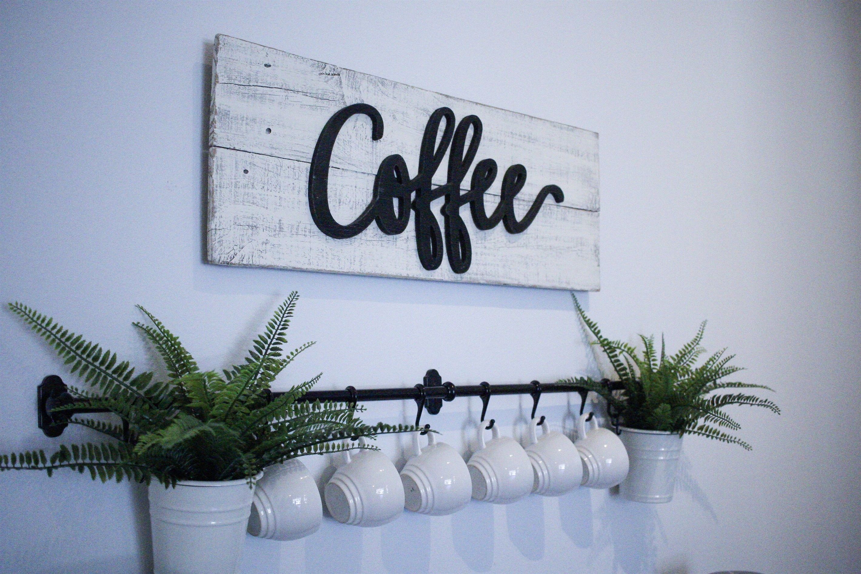 A wall-mounted sign with the word "Coffee" in black cursive letters, a black metal rack with white ceramic mugs, and two potted ferns.