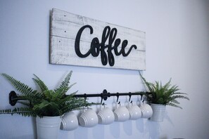 A wall-mounted sign with the word "Coffee" in black cursive letters, a black metal rack with white ceramic mugs, and two potted ferns. - Hibiscus Hideaway: Pool, Spa & Steps to the beach (Cocoa Beach)