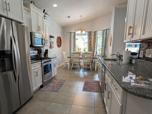 A kitchen with white cabinetry, stainless steel appliances, a dining area with a table and chairs, and a view through the window. - Hibiscus Hideaway: Pool, Spa & Steps to the beach (Cocoa Beach)