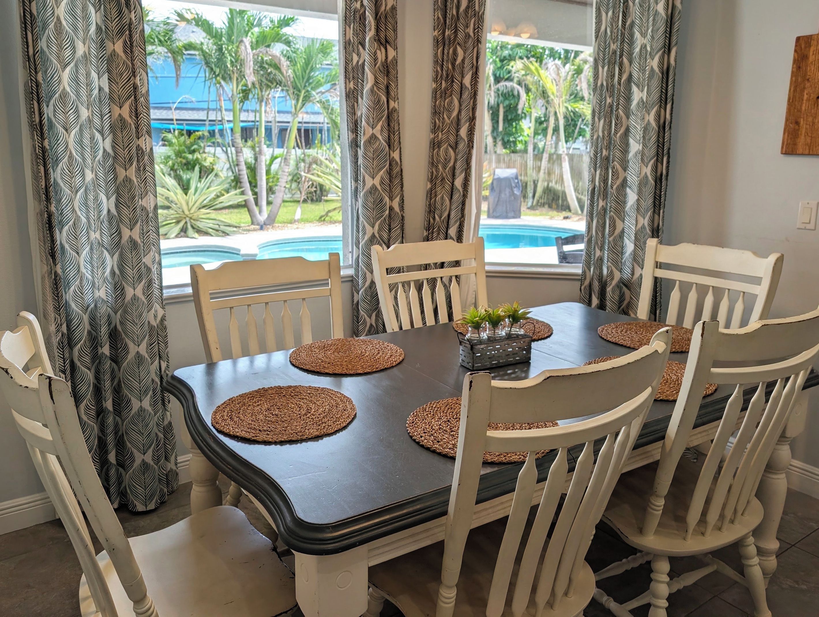 A dining table with six chairs, a central decorative item, and a view of a swimming pool and palm trees through the window.