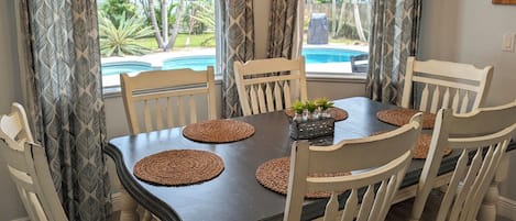 A dining table with six chairs, a central decorative item, and a view of a swimming pool and palm trees through the window.