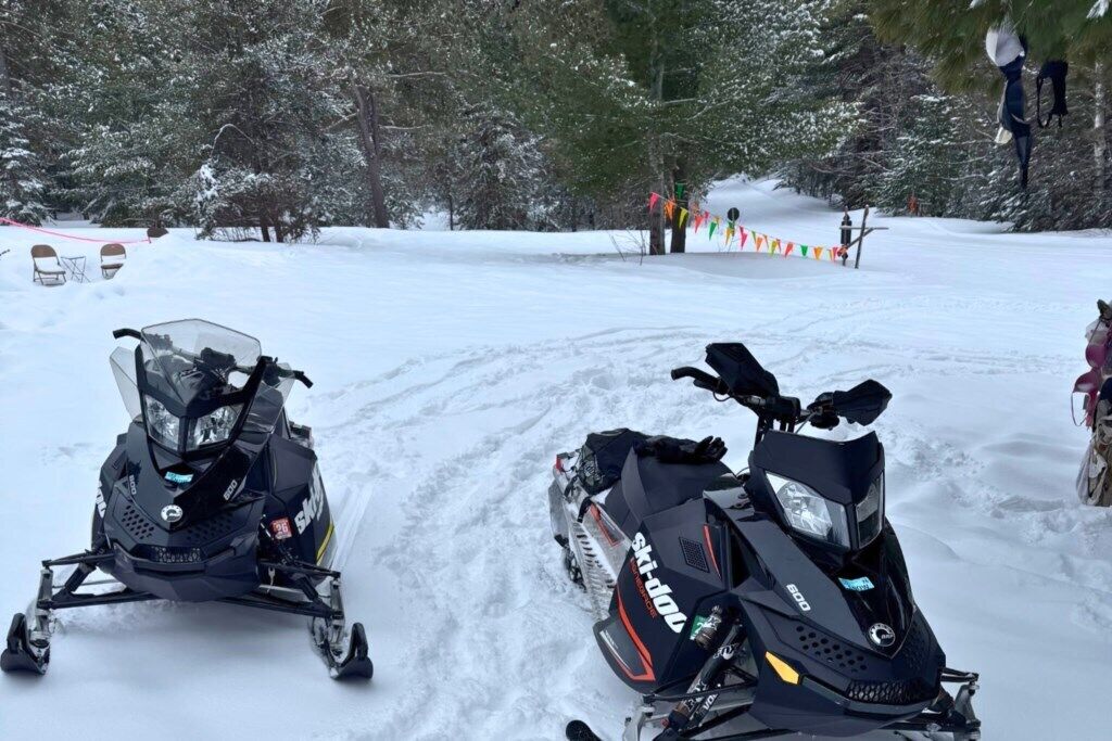 Dos motos de nieve estacionadas en un camino cubierto de nieve, con árboles al fondo.
