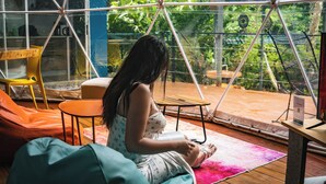 A woman reading in a room with a large window, a wooden floor, and a chair. - Bitzu Dome - Bijagua Riverside Glamping (Bijagua de Upala)