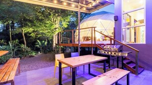 An outdoor patio with wooden tables and chairs, a barbecue grill, and a tent in the background.