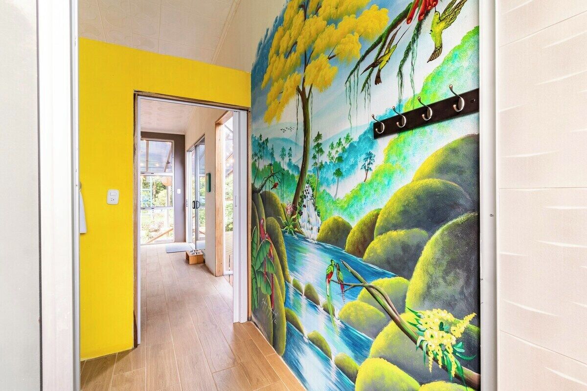 Een gang met een muurschildering van een rivier en vogels, een gele wand en een deur die leidt naar een andere kamer.