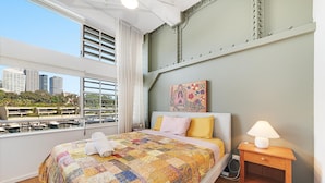 Ein Hotelzimmer mit einem großen Bett, einem Nachttisch, einer Lampe, einem Gemälde und Blick auf die Stadt durch große Fenster.
