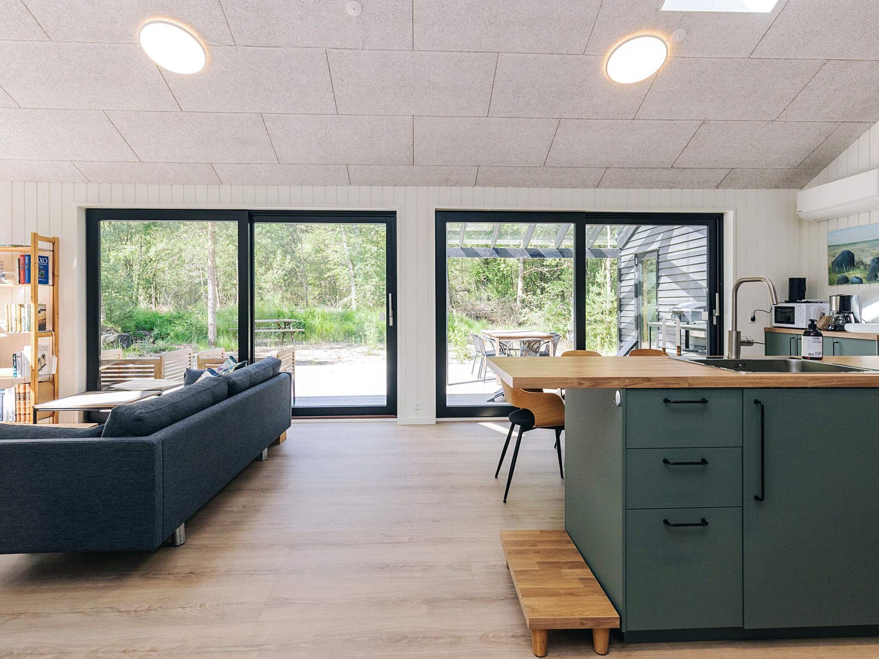 Cozinha moderna com ilha de madeira, área de refeições com mesa e cadeiras, e um sofá em sala com vista para o exterior.