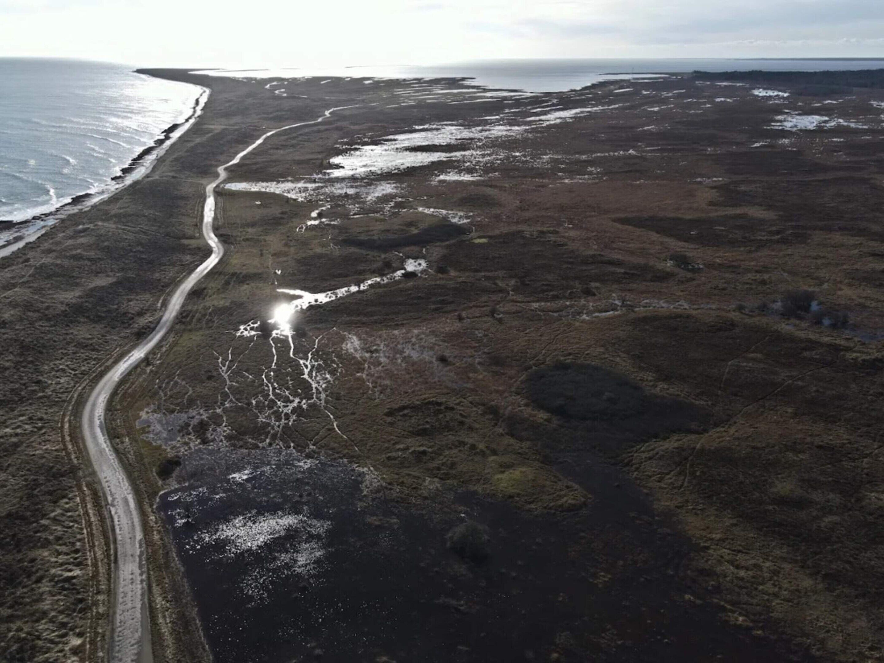Vue aérienne d’un paysage côtier, avec une route sinueuse, des zones enneigées et une ligne de côte lointaine.