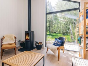 A modern living room with a wood-burning stove, two chairs, a table, and a large window overlooking a forest.