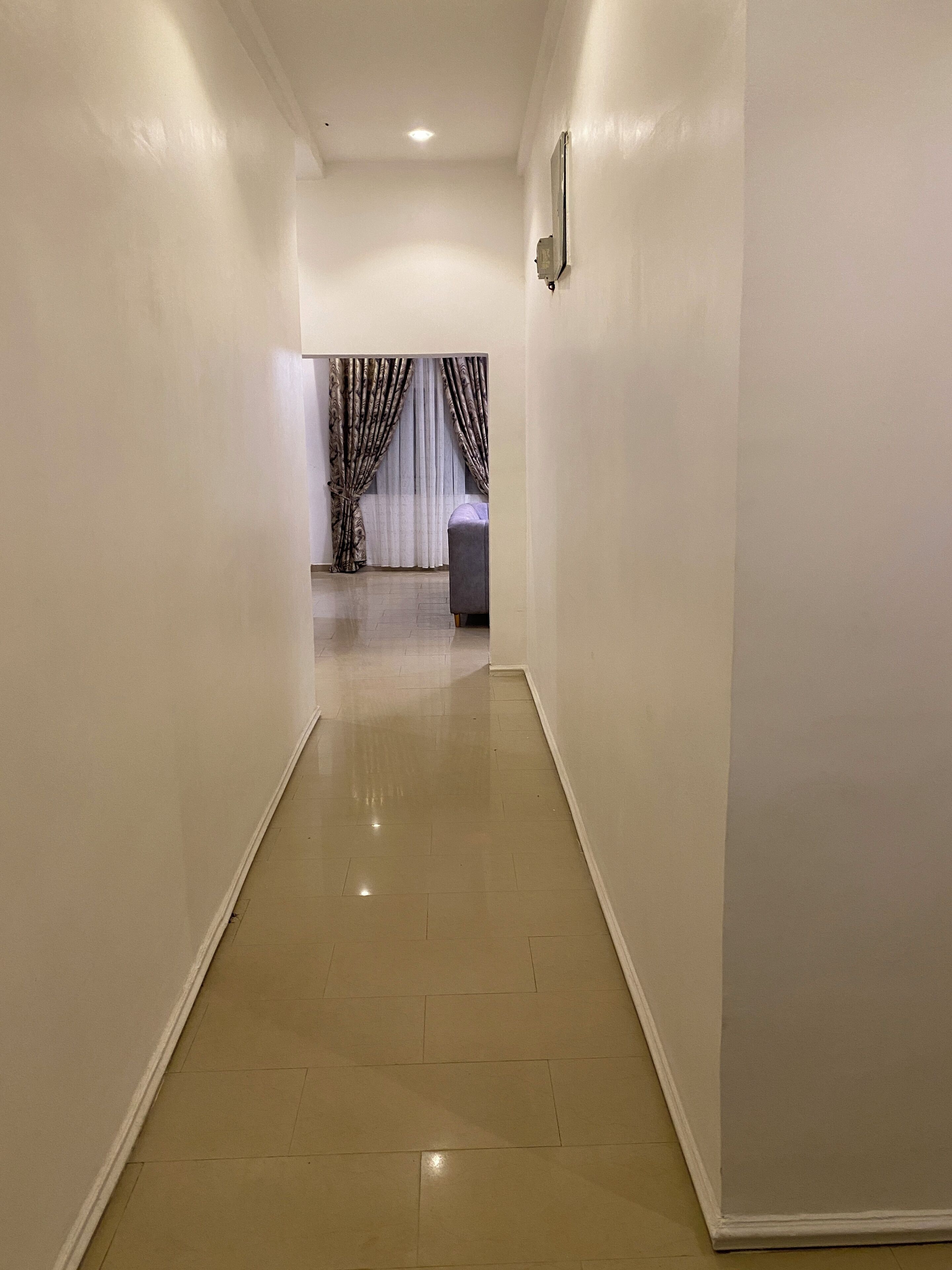 A hallway with white walls and a shiny tiled floor, leading to a room with a bed and curtains.