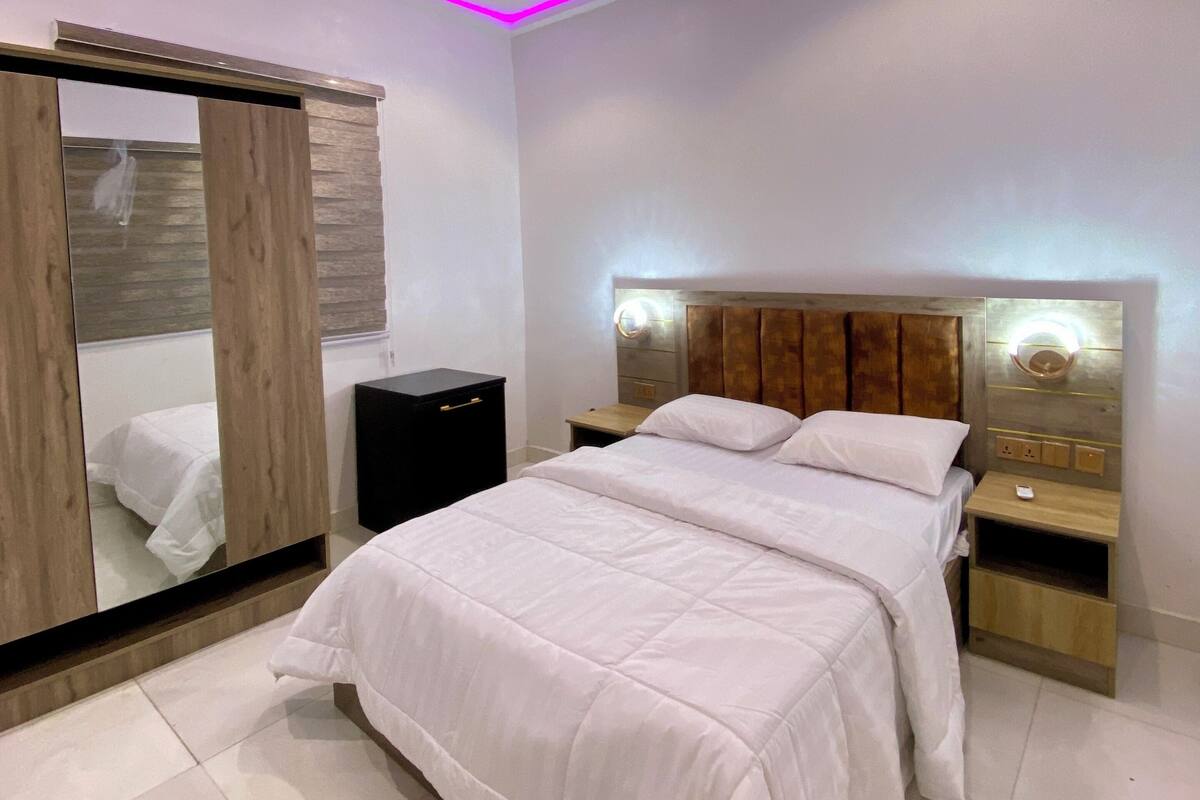 Ein modernes Schlafzimmer mit einem großen Bett, einem Schrank mit Spiegel, Nachttischen und einer Decke mit rosafarbenen LED-Leuchten.