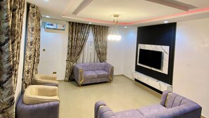A modern living room with a large flat-screen TV mounted on a marble wall, a sofa set, and a ceiling with recessed lighting and red LED strip lighting. - BA2 Homes (Abuja)